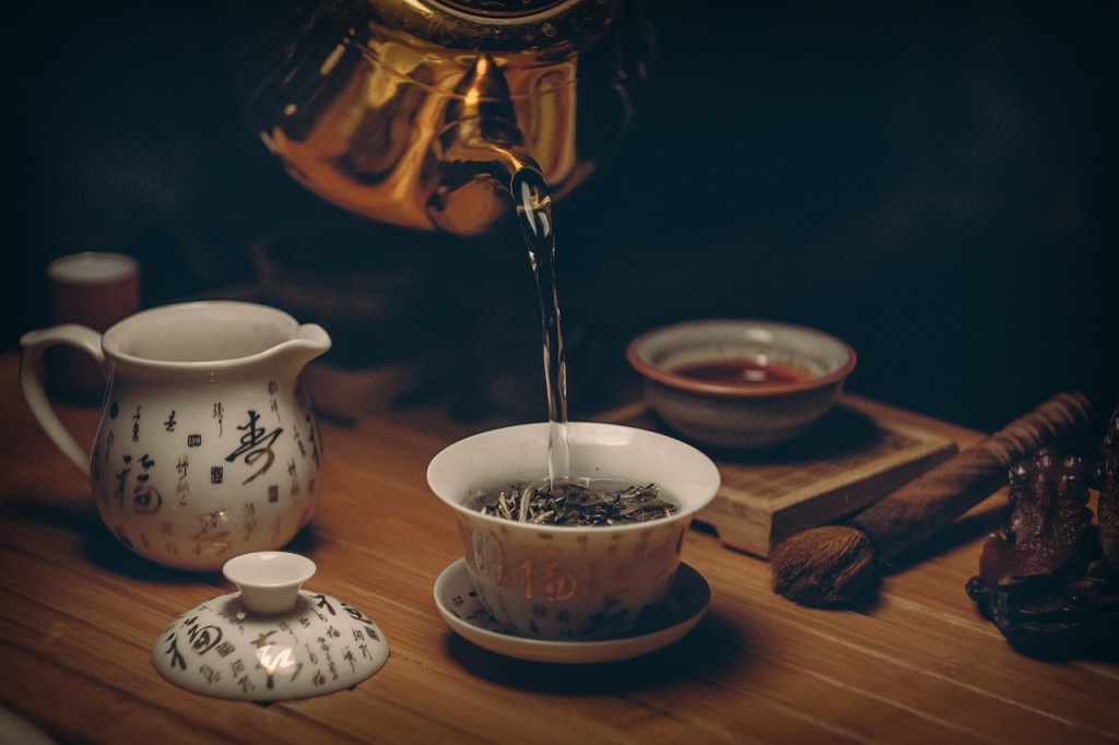 A kettle pouring water into a tea cup with tea leaves in it.